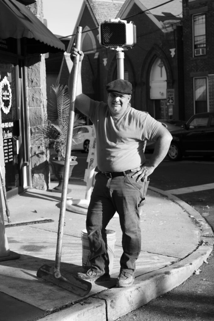 Black and white photograph of a middle-aged man standing with a long pole-like tool, on a street corner.