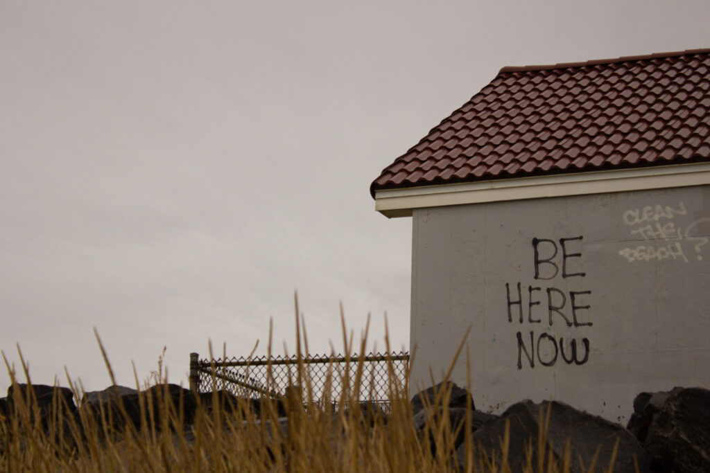 Photograph of the side of a building with graffiti saying "be here now" and "clean the beach."