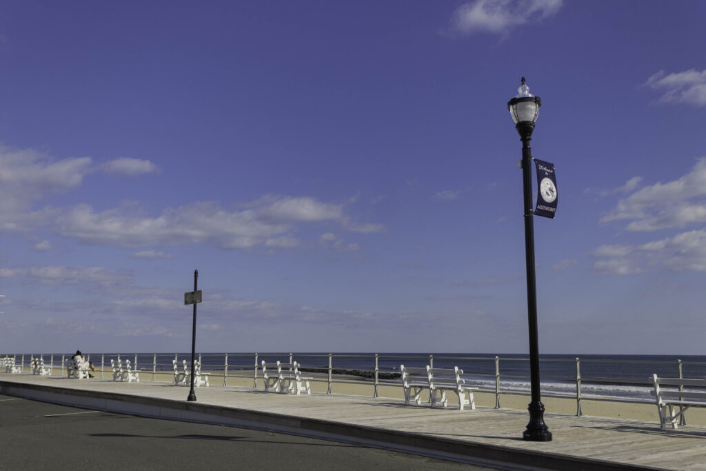 Photograph of a boardwalk on the beach, with some white benches and a lampost. Most of the frame shows a blue sky with a few clouds.