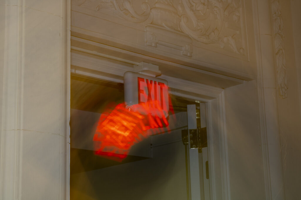 Photography of an exit sign. The word "EXIT" appears to be multiplied and falling out of the sign.